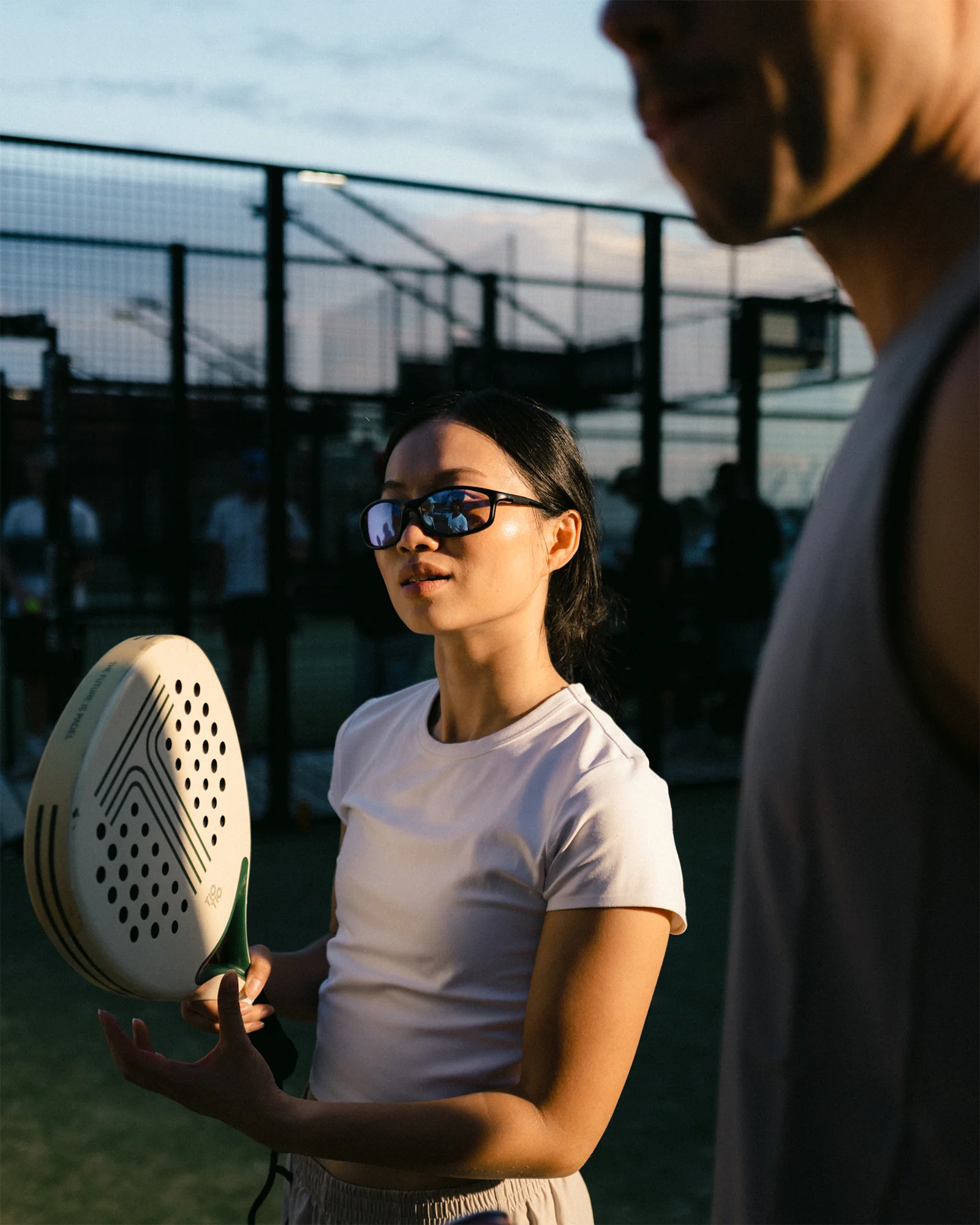 Sportliche Frau hält Padel vor abendlichem Hintergrund mit Person in der Nähe